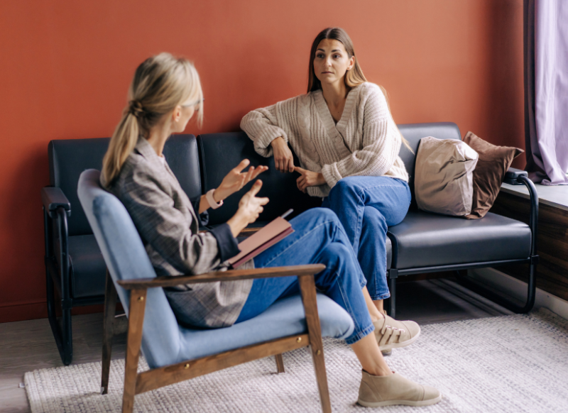 Two women engaged in conversation while sitting on a couch, displaying a friendly and relaxed atmosphere.