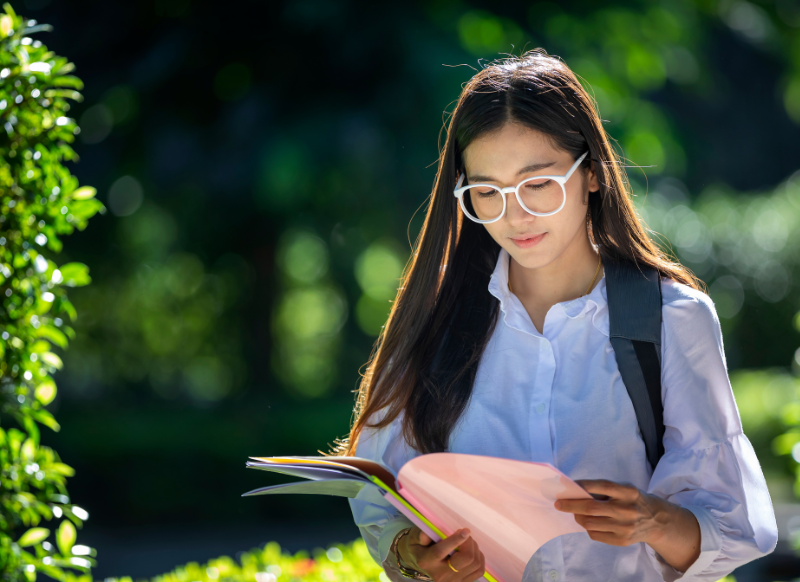 student sitting on a bench in a park, focused on reading a book with trees and grass in the background.