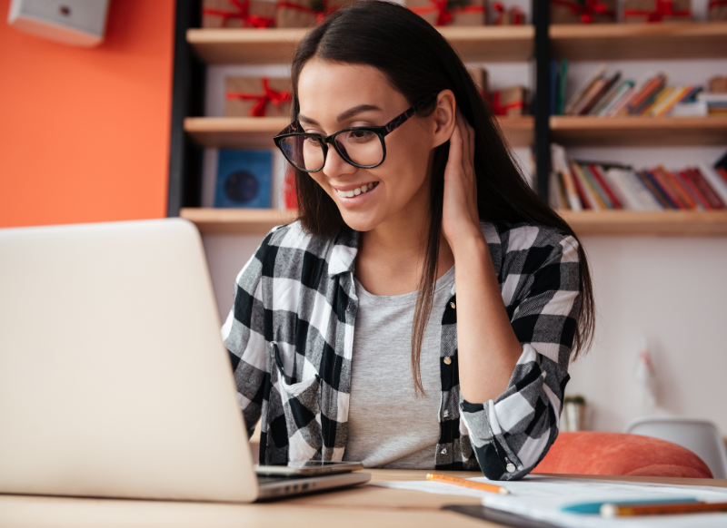 A woman wearing glasses smiles while working on her laptop, showcasing a positive and engaged expression.