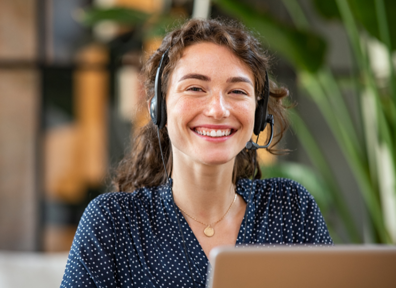 A woman with a headset smiles while working on a laptop, conveying a sense of engagement and positivity.